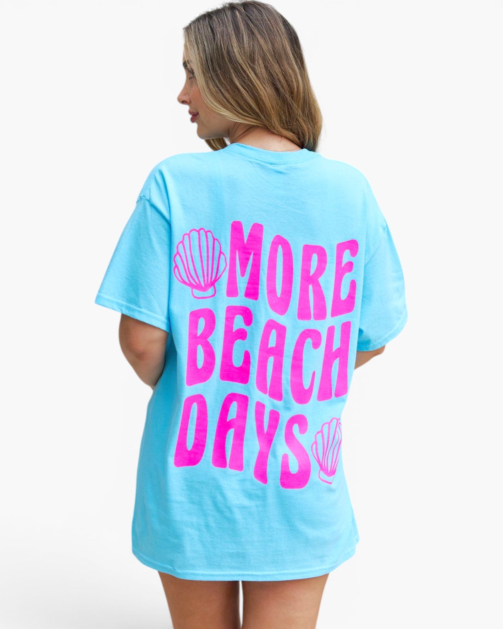 A woman shown from the back wearing a light blue oversized t-shirt with a bright pink graphic that reads 'MORE BEACH DAYS' with seashell illustrations, set against a plain white background.