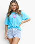 A woman modeling a light blue oversized t-shirt with a pink seashell graphic on the chest, paired with light-wash denim shorts, against a plain white background.