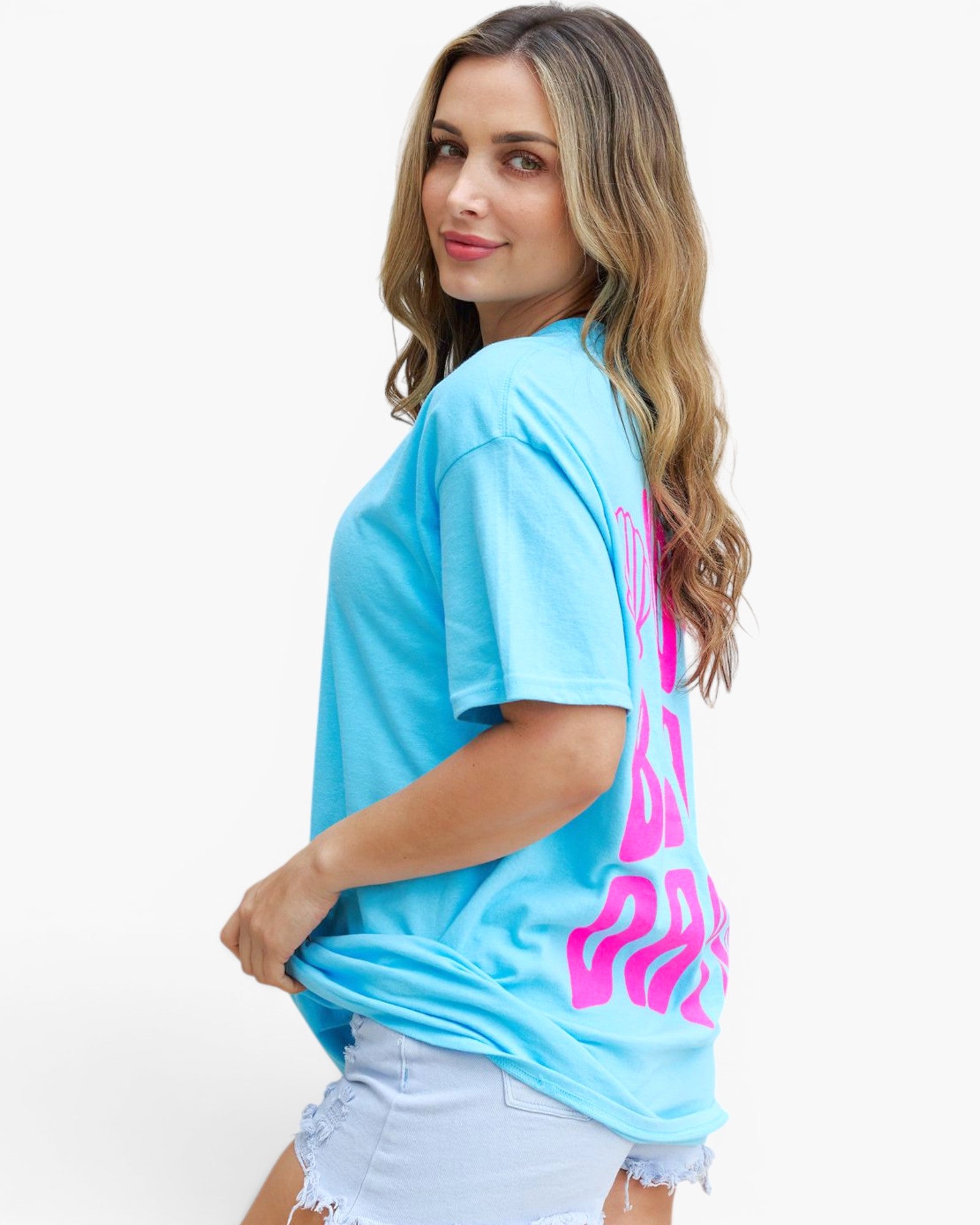 A woman wearing a bright light blue oversized t-shirt with a large pink graphic print on the back, paired with light denim shorts, photographed against a solid white background.