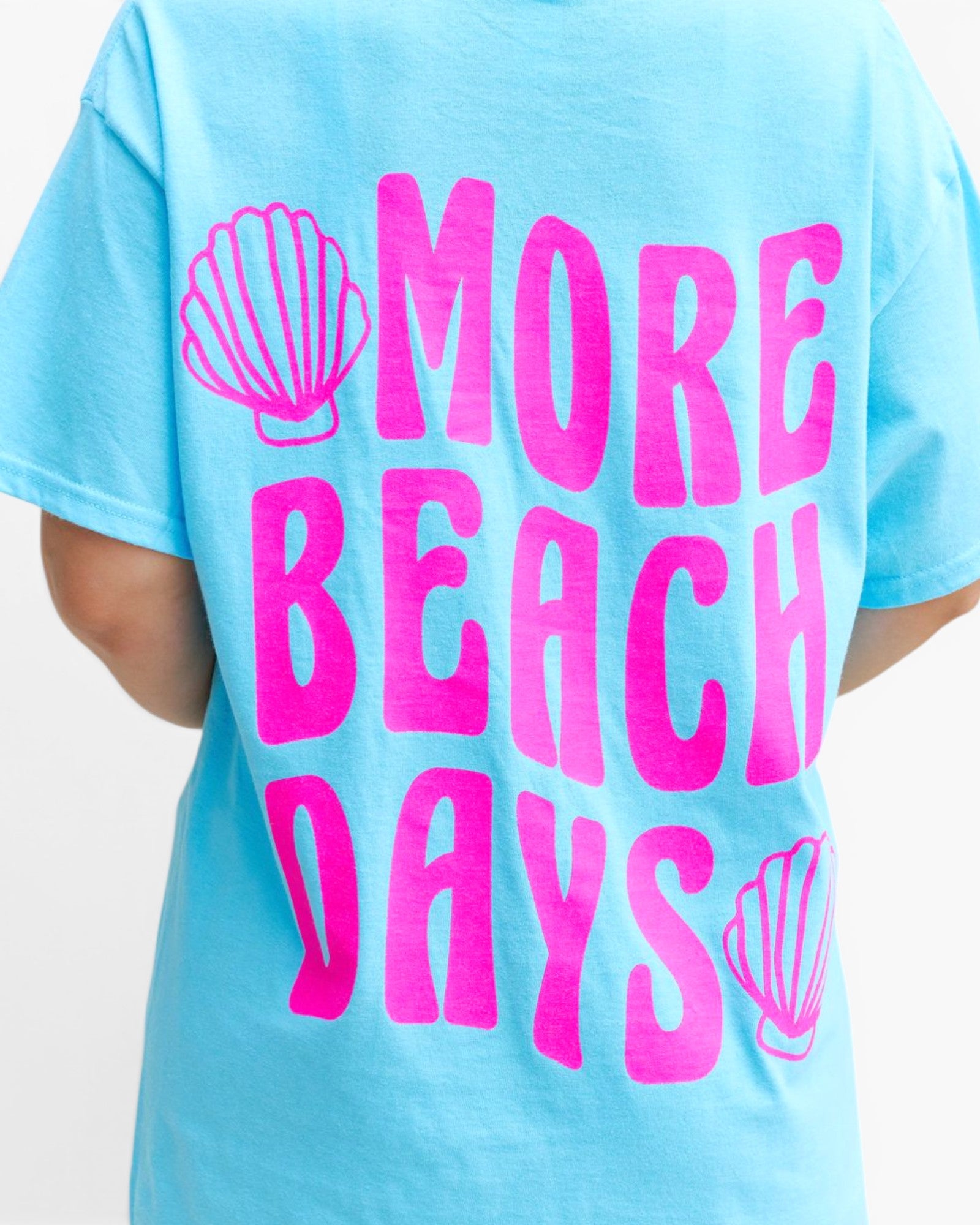 A light blue short-sleeve t-shirt with a bright pink graphic print that reads 'MORE BEACH DAYS' accompanied by two seashell illustrations, shown on a model from the back.