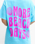 A light blue short-sleeve t-shirt with a bright pink graphic print that reads 'MORE BEACH DAYS' accompanied by two seashell illustrations, shown on a model from the back.