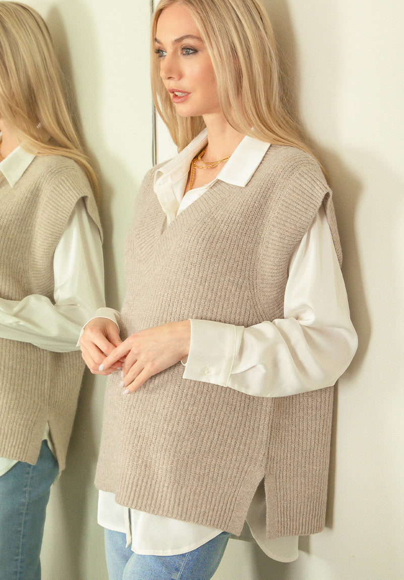 Woman wearing a beige knitted vest over a white shirt and blue jeans against a neutral background.