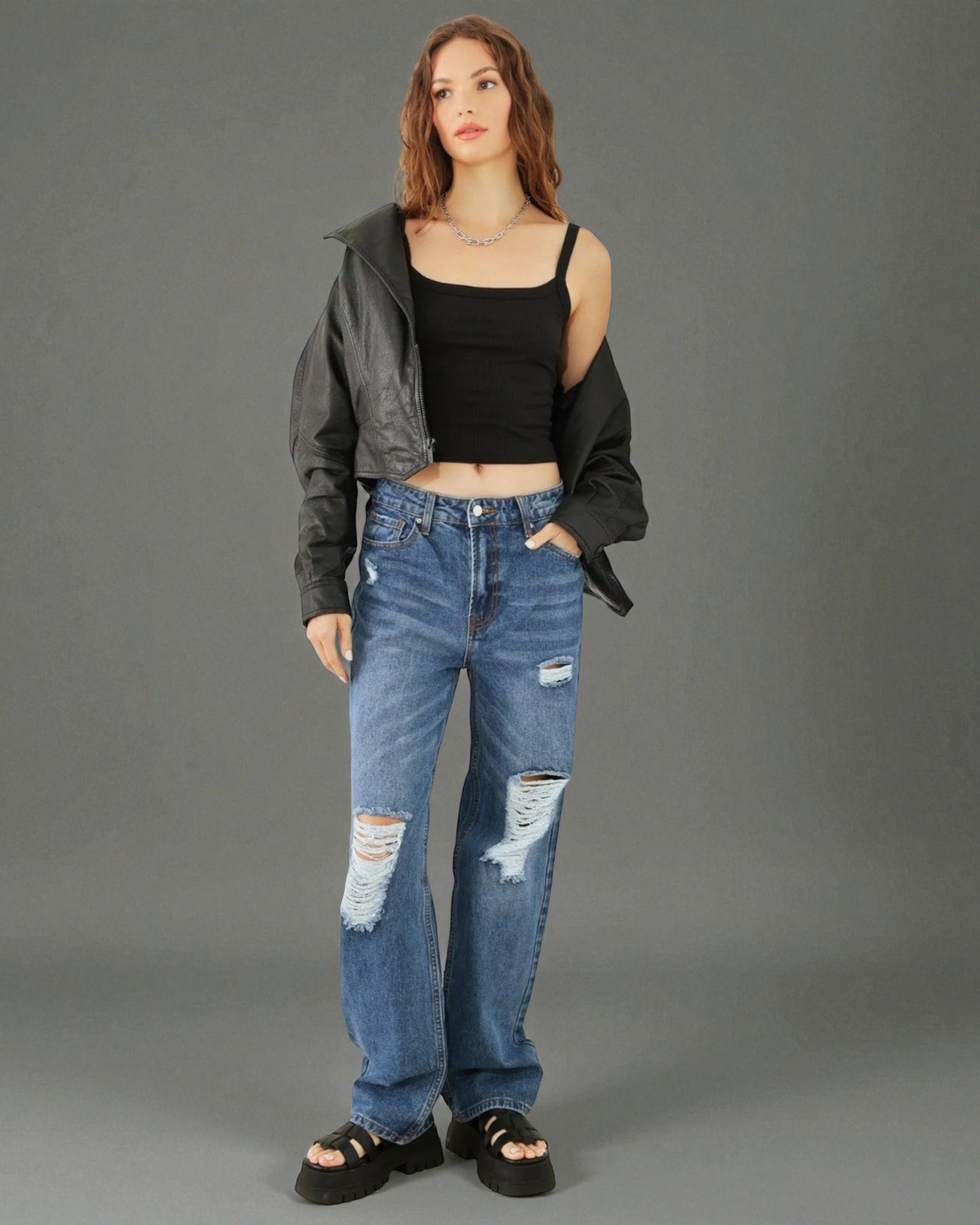 Woman wearing a black crop top, gray jacket, and blue ripped jeans on a gray background
