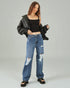 Woman wearing a black leather jacket, black crop top, and blue distressed jeans on a gray background