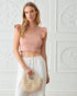 Woman wearing a pink top and white pants holding a straw bag against a white wall.