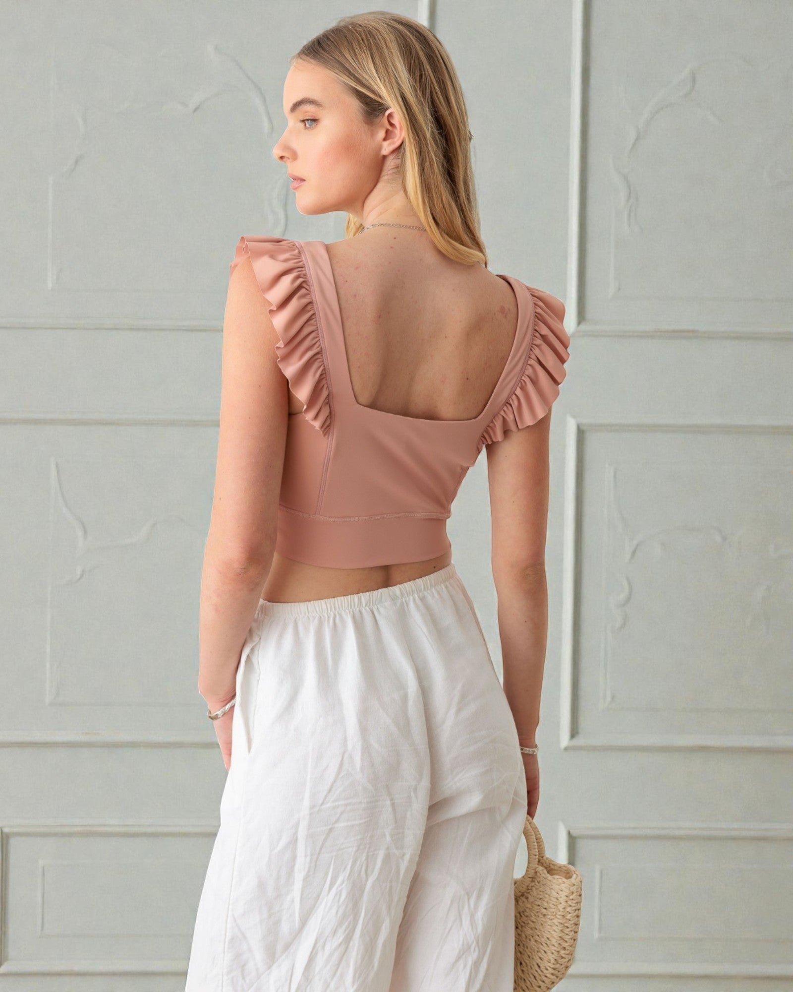 Woman wearing a pink crop top with ruffled shoulders and white pants against a light gray wall.