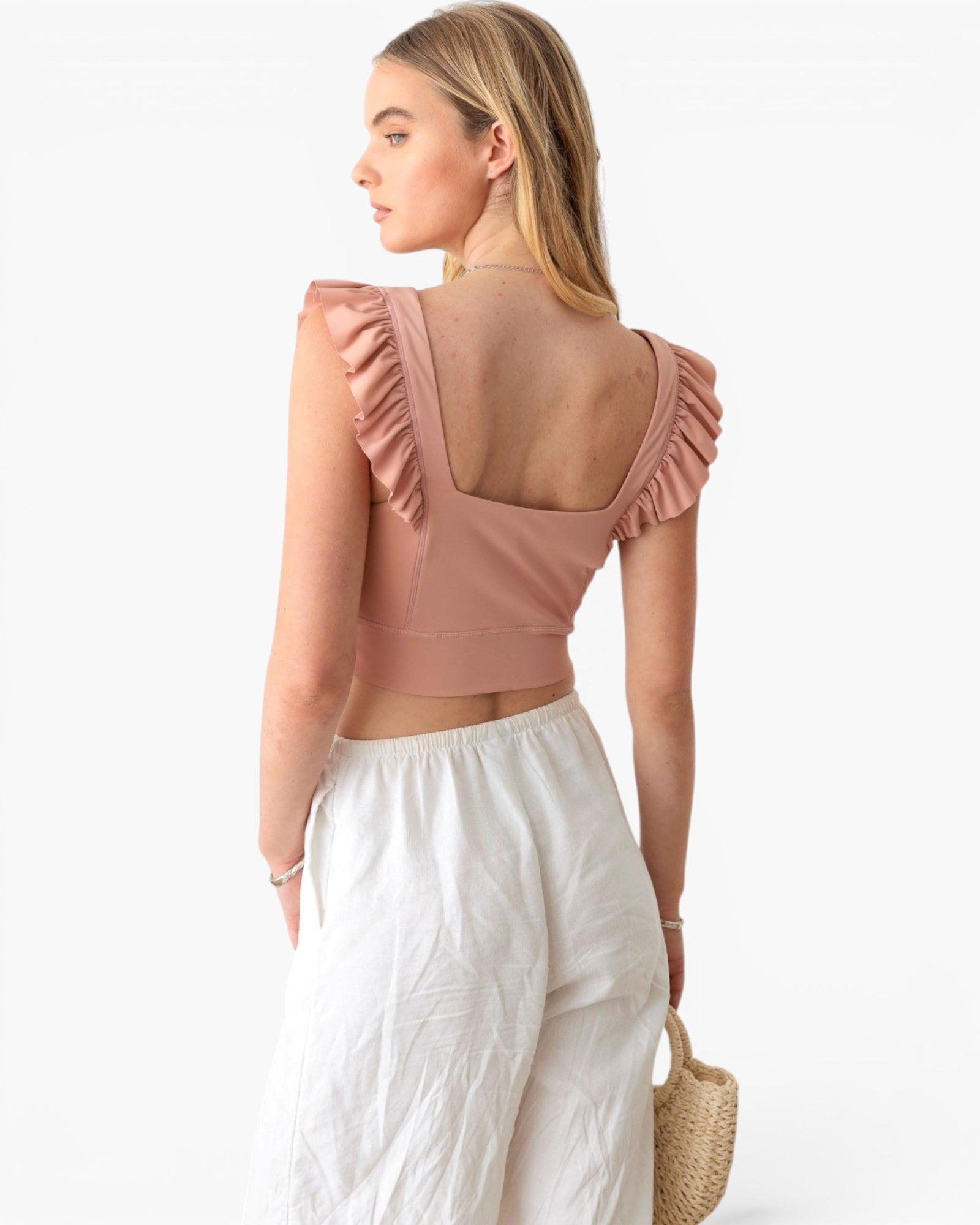 A woman shown from the back wearing a dusty rose ruffled crop top and white wide-leg pants, holding a woven straw bag against a plain white background.