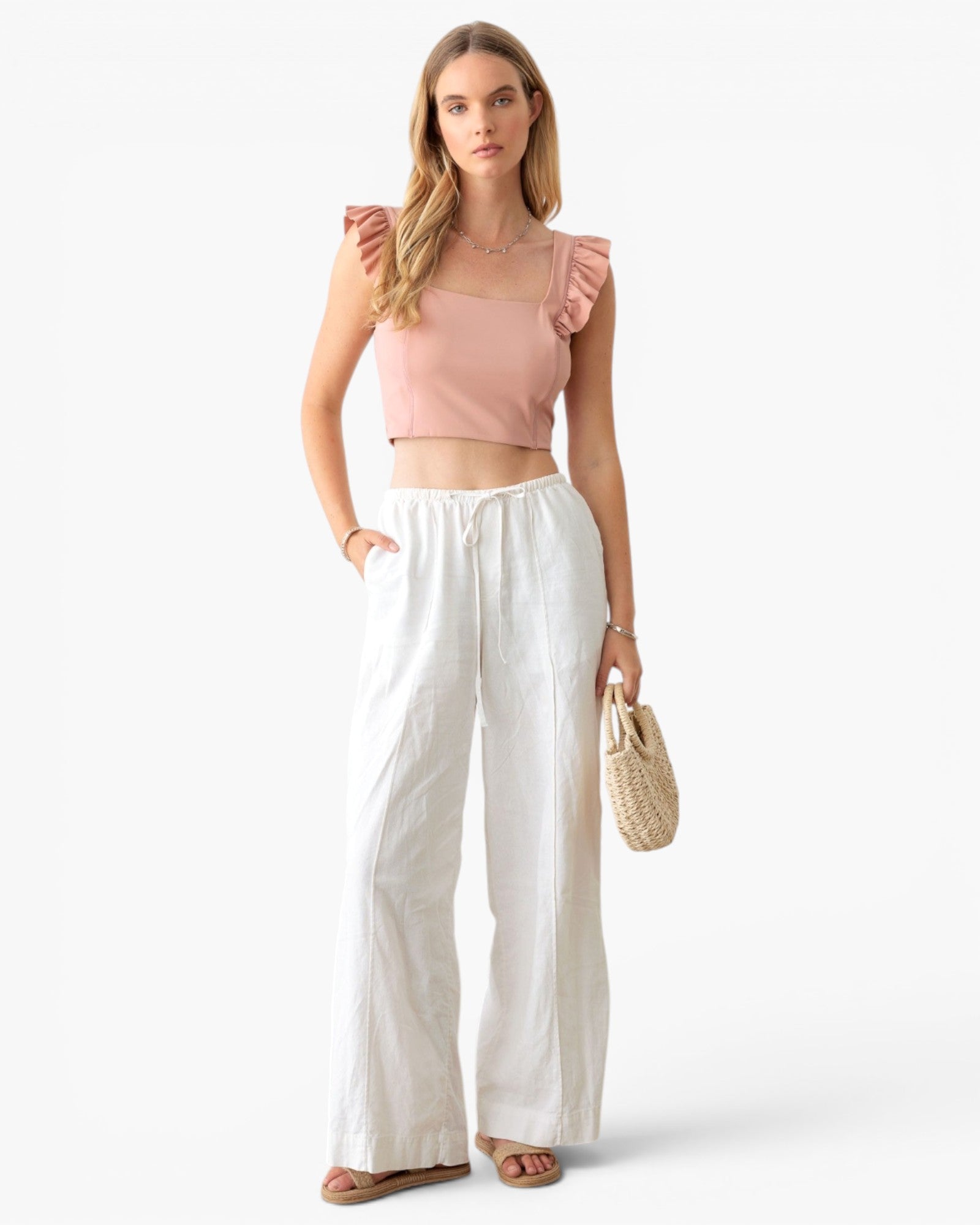 A woman modeling a light pink ruffle-shoulder crop top and white wide-leg pants, holding a small woven straw bag against a clean white studio background.
