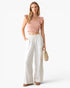 A woman modeling a light pink ruffle-shoulder crop top and white wide-leg pants, holding a small woven straw bag against a clean white studio background.