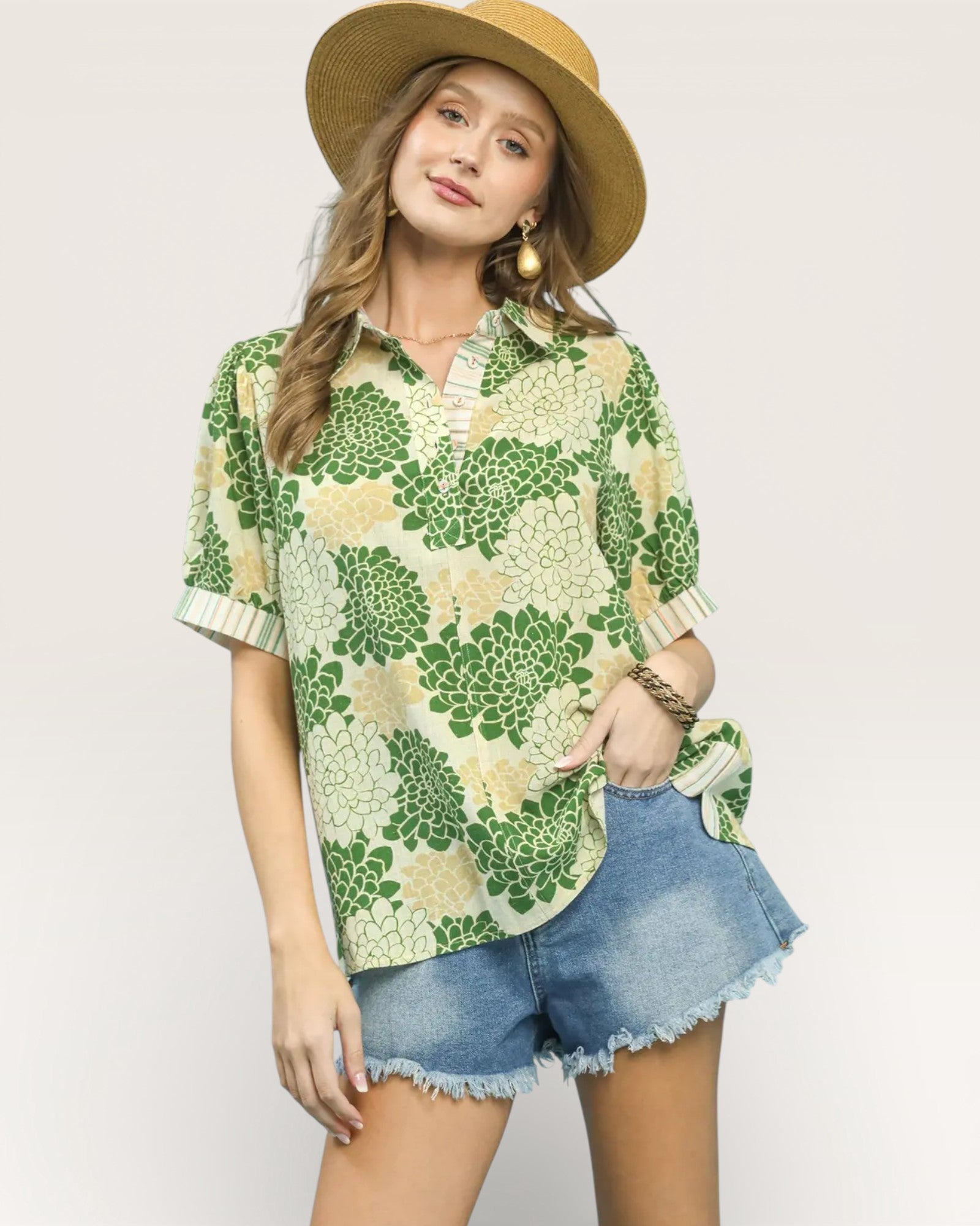 Woman wearing a green floral shirt and denim shorts with a beige hat on a plain background