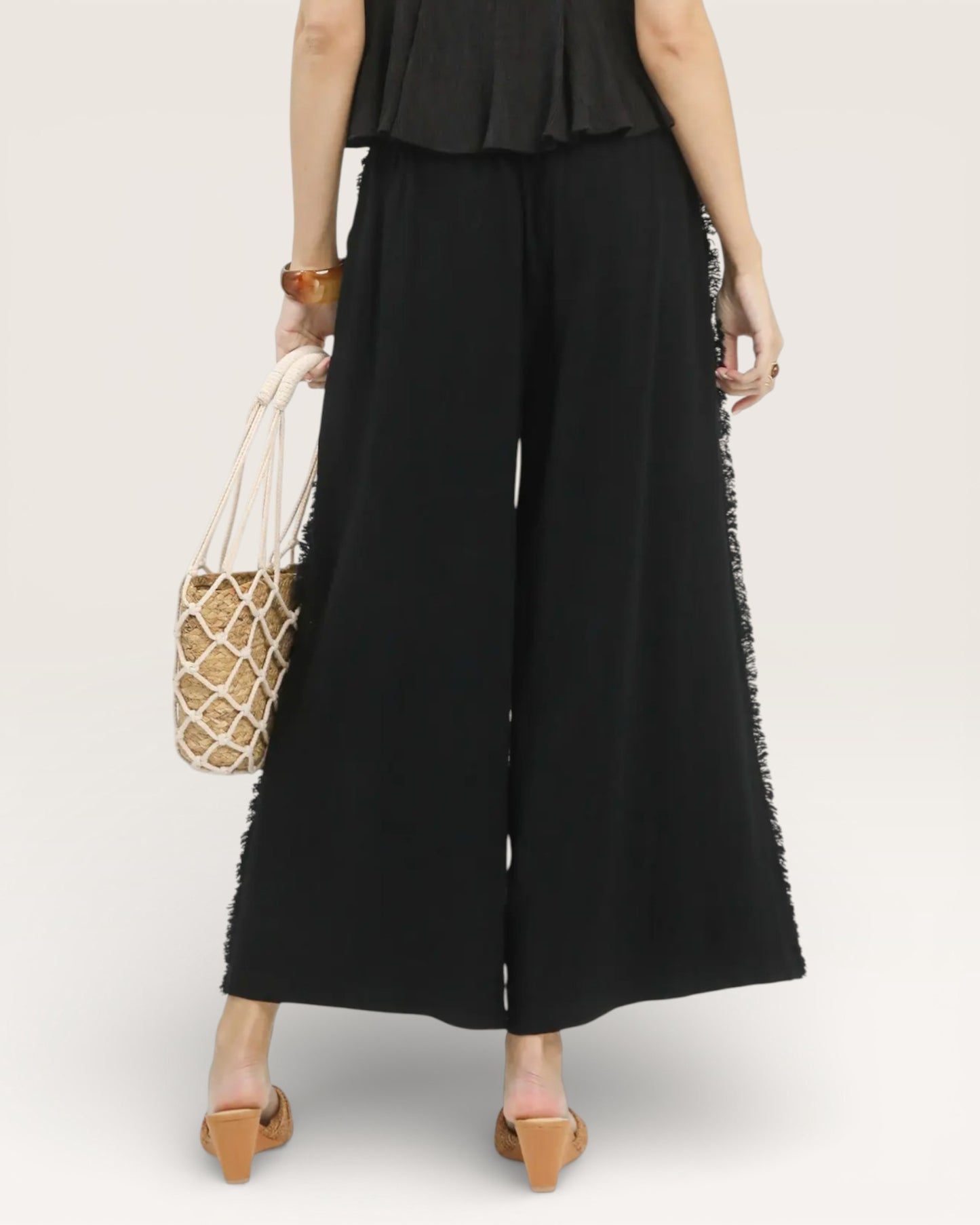 Person wearing a black outfit with wide-leg pants and a ruffled top, holding a straw bag on a white background.