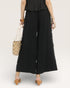 Person wearing a black outfit with wide-leg pants and a ruffled top, holding a straw bag on a white background.