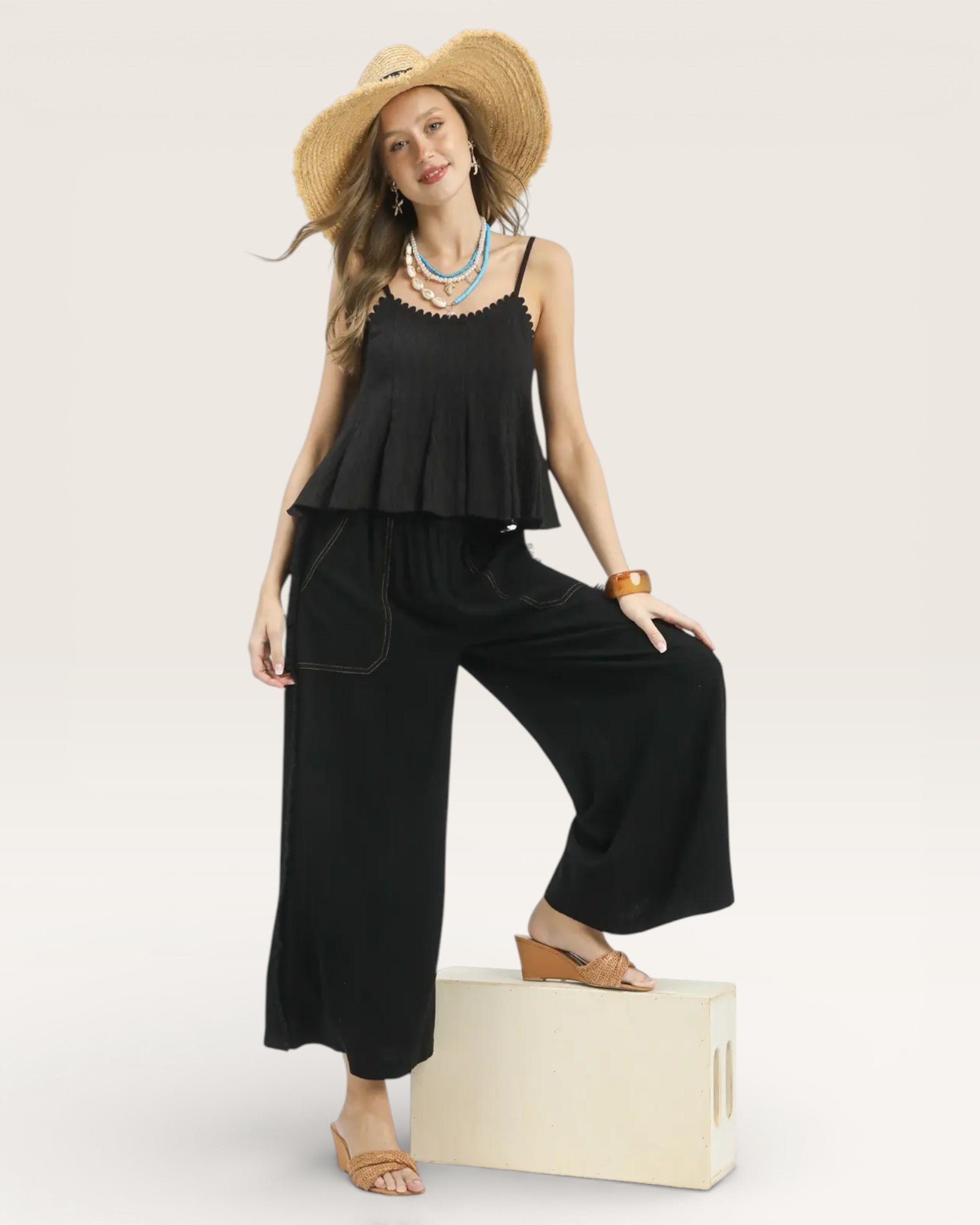 Woman wearing a black outfit with wide-leg pants and a straw hat on a white background