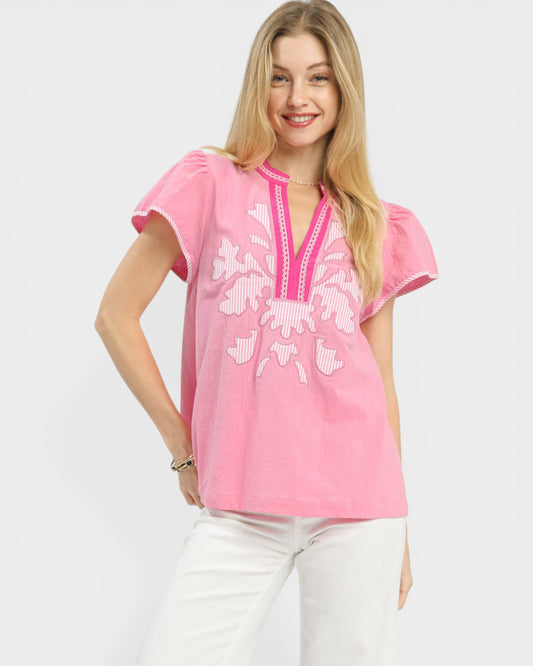 Woman wearing a pink embroidered top with white pants on a plain background