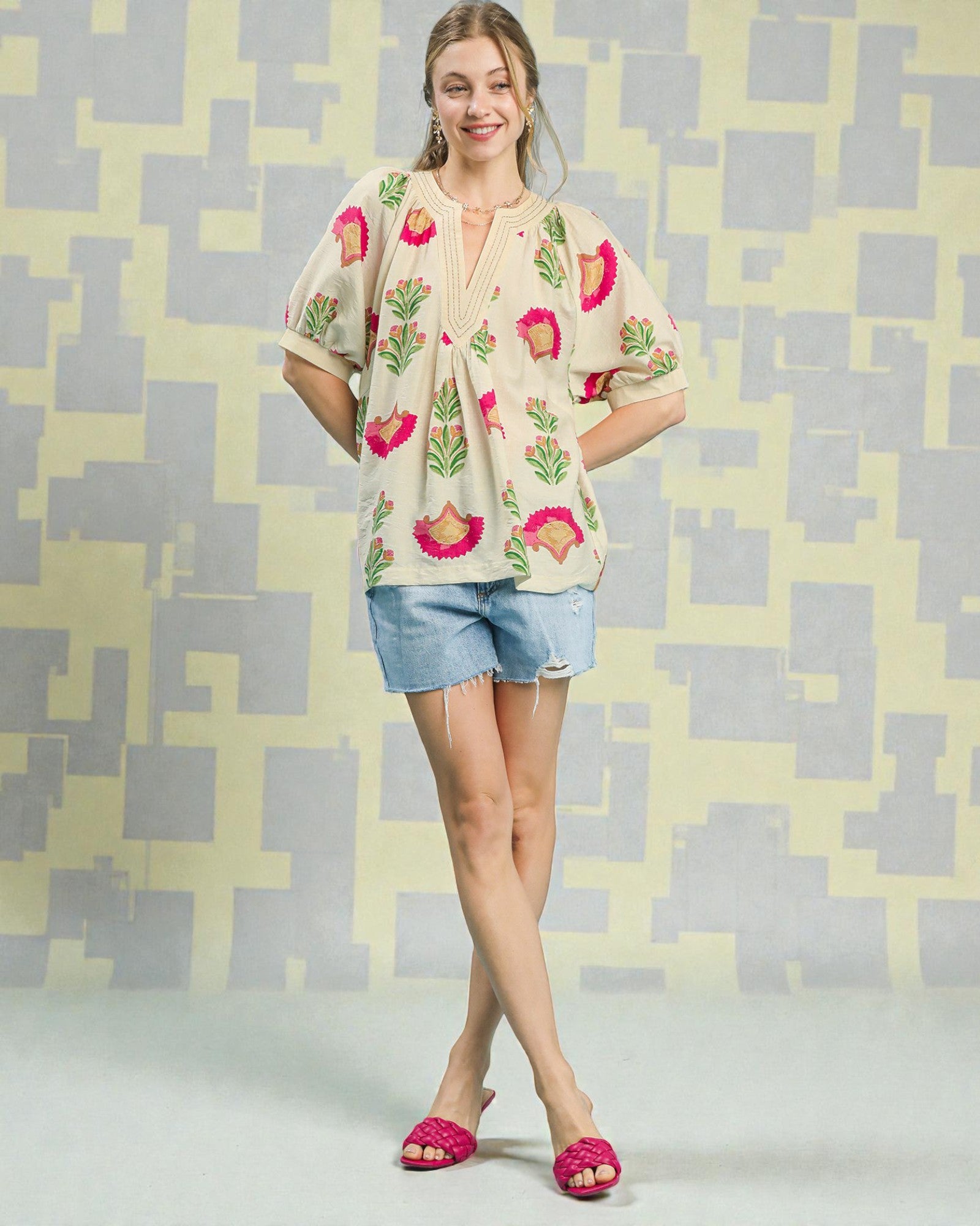 Woman wearing a floral blouse and denim shorts against a geometric patterned wall.