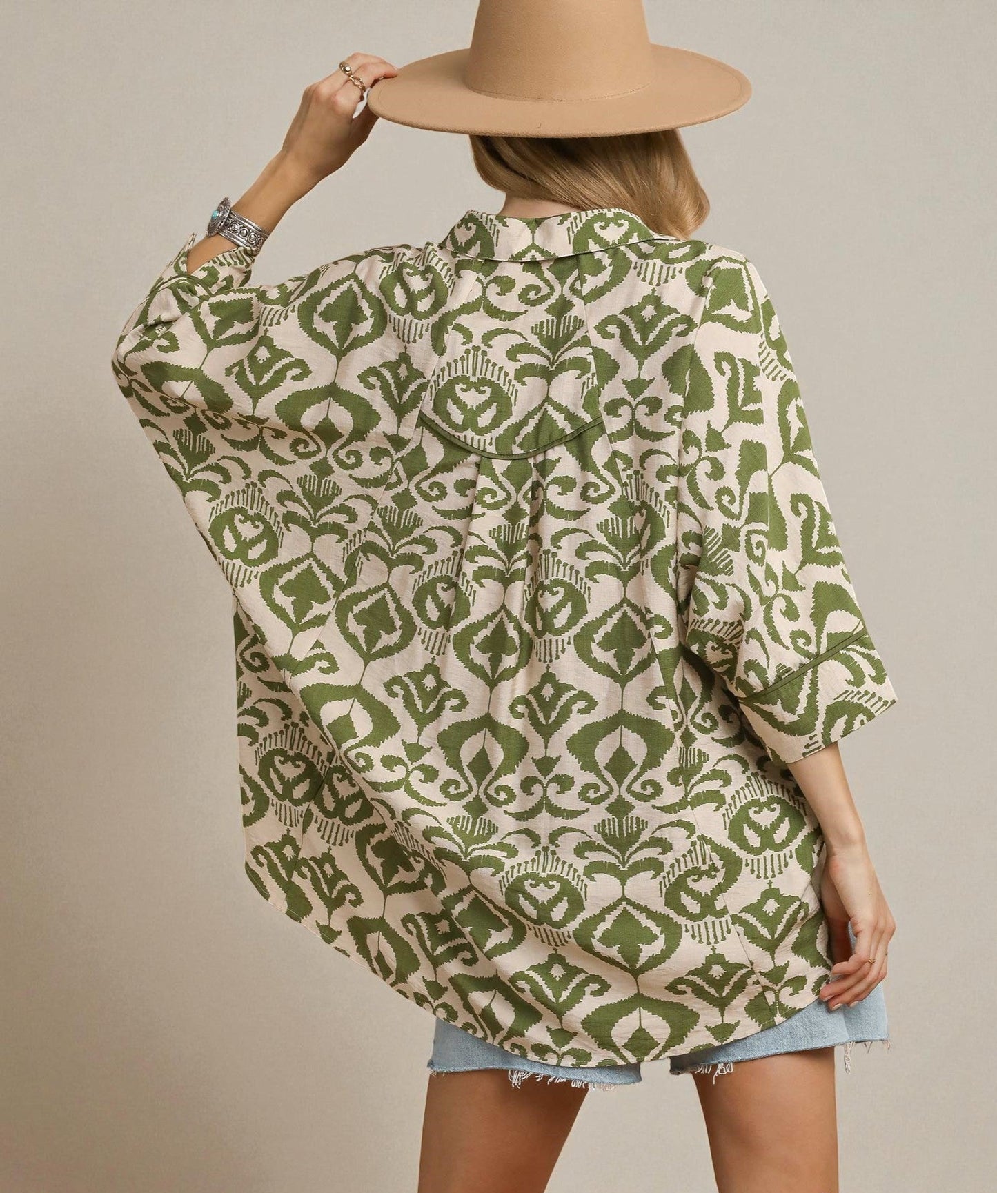 Person wearing a green and beige patterned shirt with a beige hat on a plain background