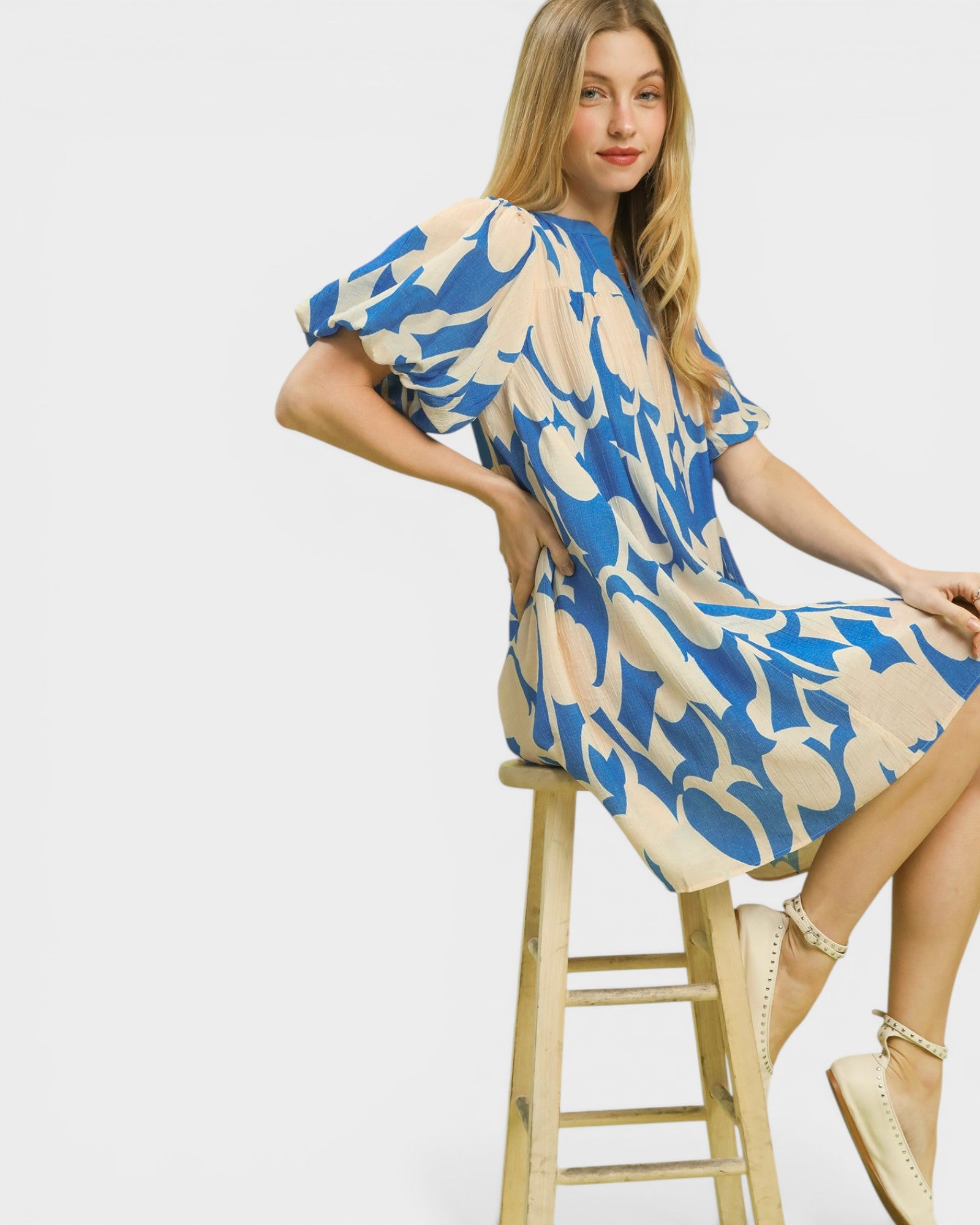 Woman sitting on a stool wearing a blue and beige patterned dress against a white background