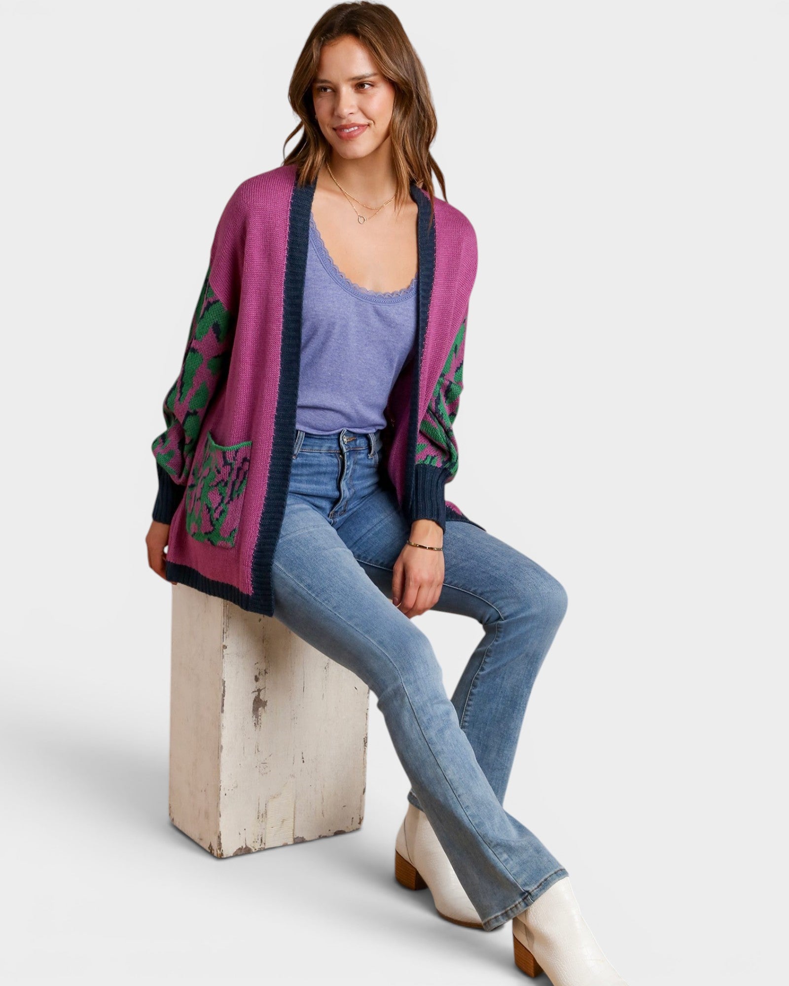 Woman wearing a purple cardigan with green patterns, blue jeans, and white shoes on a light gray background.