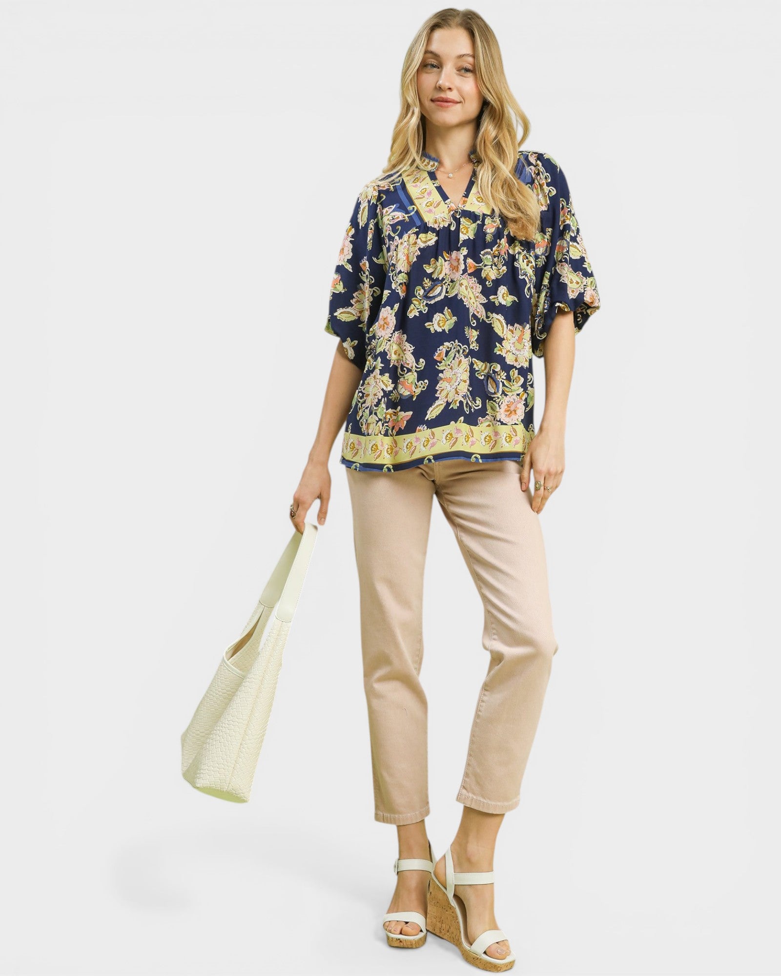 Woman wearing a floral blouse and beige pants, holding a tote bag on a white background