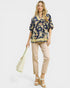 Woman wearing a floral blouse and beige pants, holding a tote bag on a white background