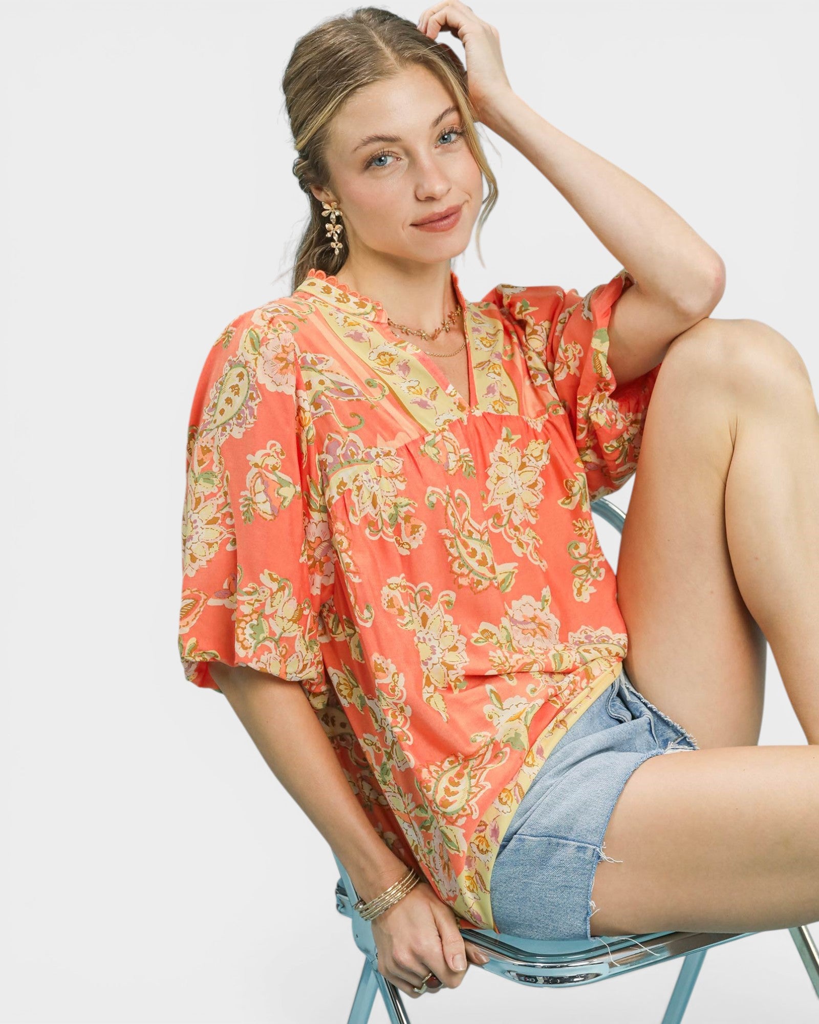 Woman wearing a floral blouse and denim shorts sitting on a chair against a plain background