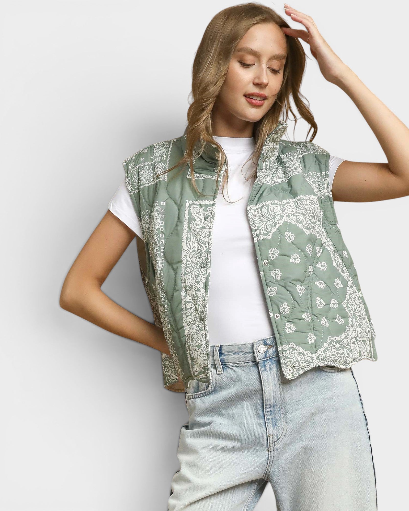 Woman wearing a green patterned vest over a white shirt and light blue jeans on a plain background