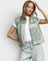 Woman wearing a green patterned vest over a white shirt and light blue jeans on a plain background