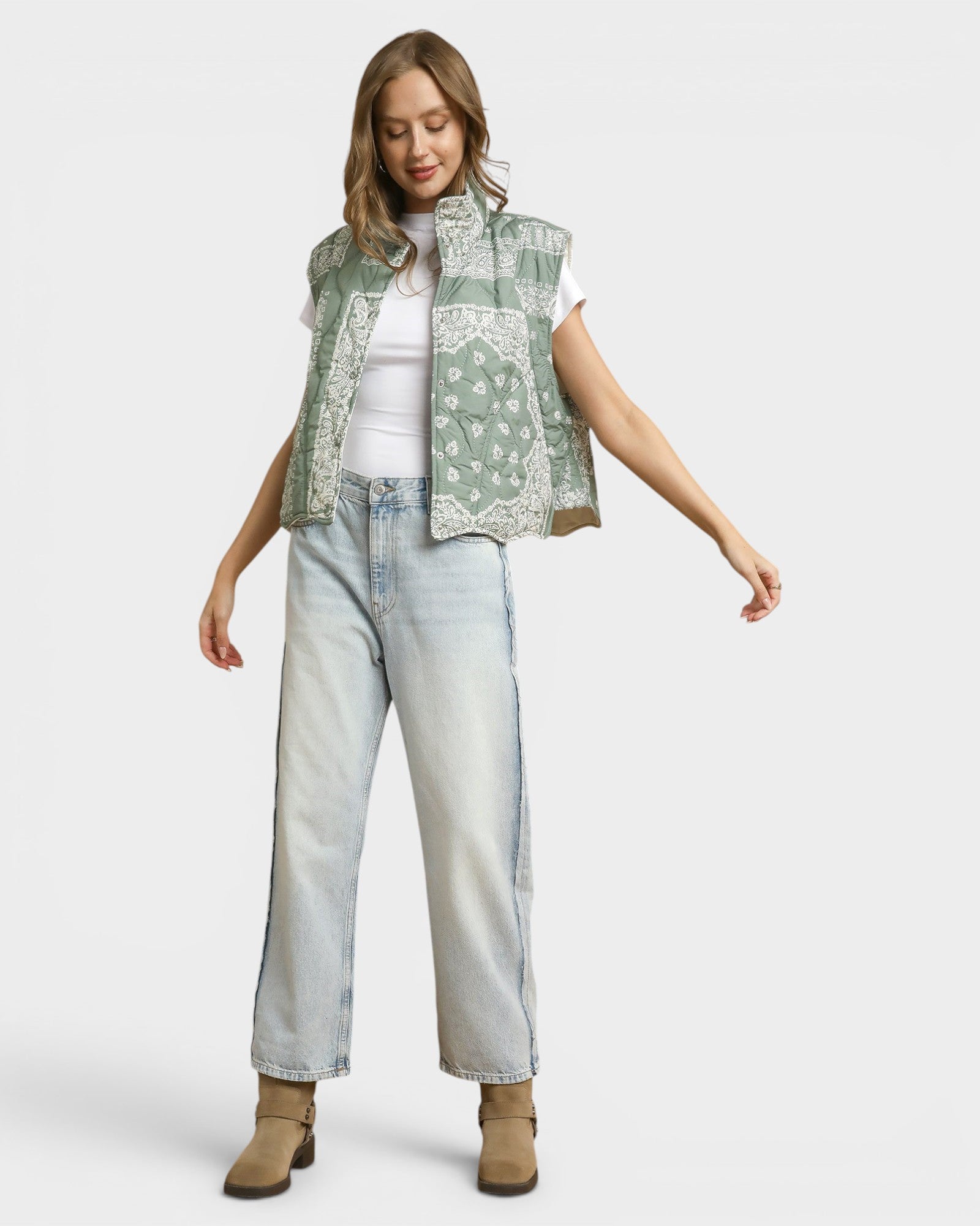 Woman wearing a green patterned vest, white shirt, light blue jeans, and brown boots on a white background