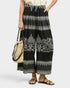 A person wearing black wide-leg cropped pants with white bohemian-style floral and geometric patterns, holding a woven straw bucket bag and wearing brown strappy sandals against a light gray background.