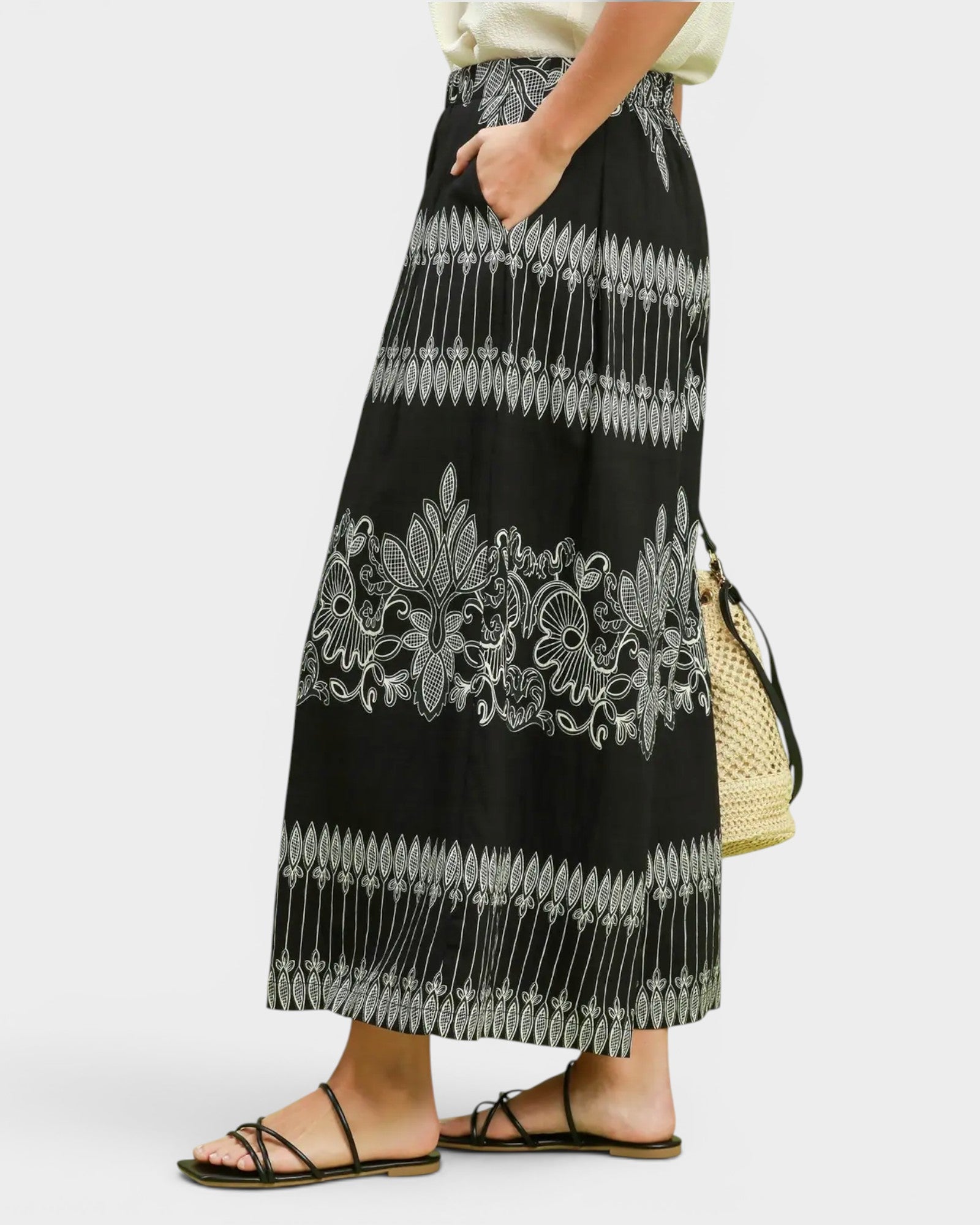 A person wearing a long black maxi skirt with white embroidered floral and geometric patterns, paired with black strappy flat sandals and holding a woven straw bag, against a light gray studio background.