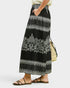 A person wearing a long black maxi skirt with white embroidered floral and geometric patterns, paired with black strappy flat sandals and holding a woven straw bag, against a light gray studio background.
