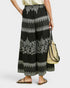 A model is shown from the waist down wearing black wide-leg cropped pants with intricate white and gray bohemian-style embroidery and patterns. The model is holding a woven straw bucket bag and wearing black strappy flat sandals against a light gray studio background.