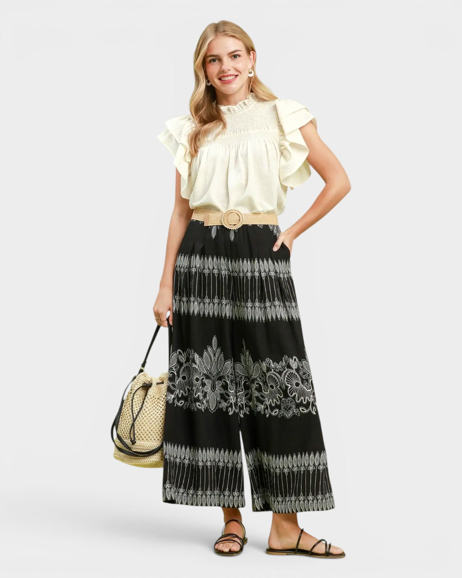 A woman modeling a cream ruffled short-sleeve blouse paired with wide-leg black pants featuring gray floral and geometric patterns, holding a woven beige handbag against a light gray studio background.