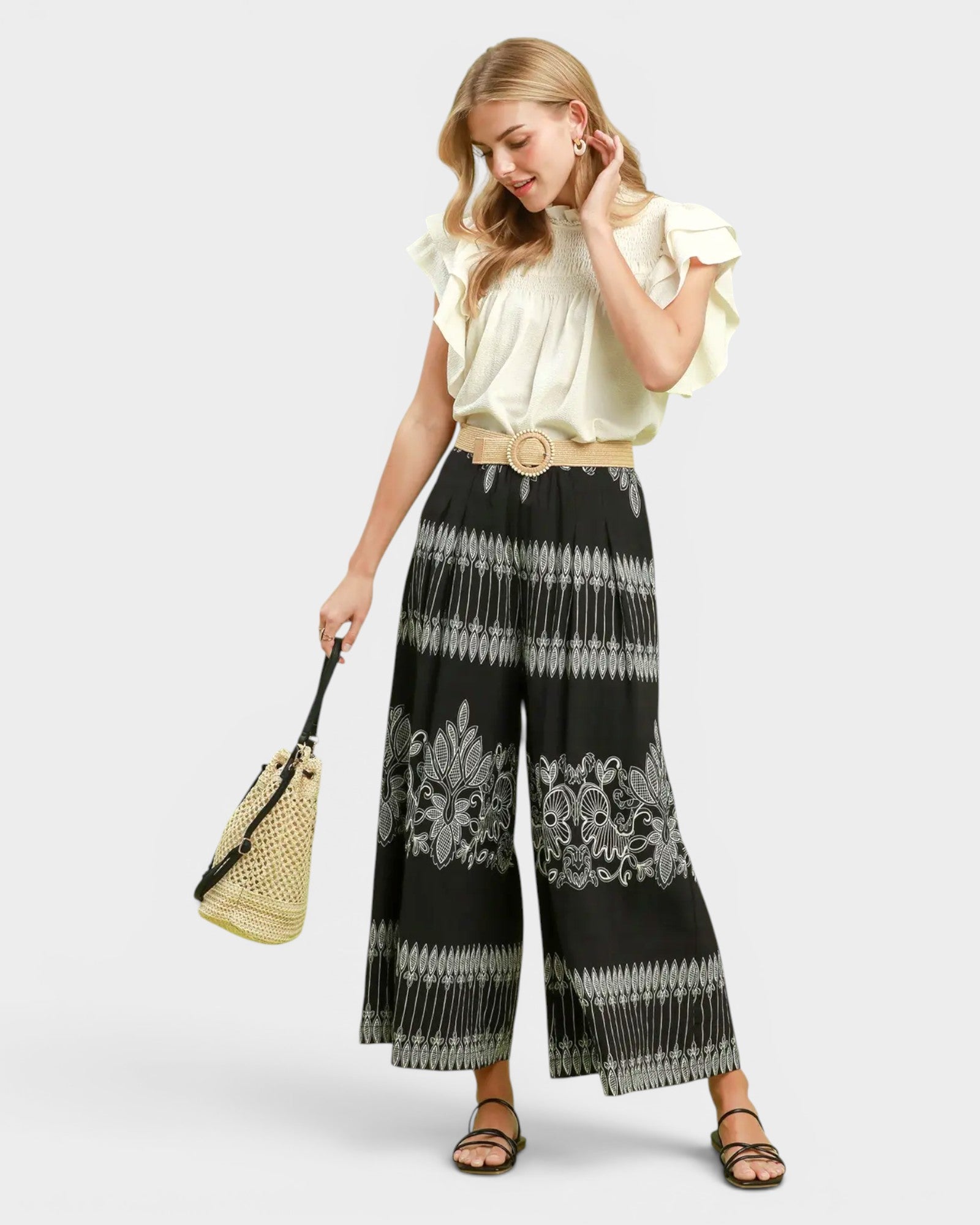 A woman modeling a cream ruffled blouse and black wide-leg pants with a white ikat pattern, styled with a tan belt, a woven straw bag, and brown sandals against a light gray background.