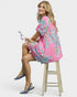 Woman wearing a pink dress with colorful patterns, sitting on a wooden stool against a white background.