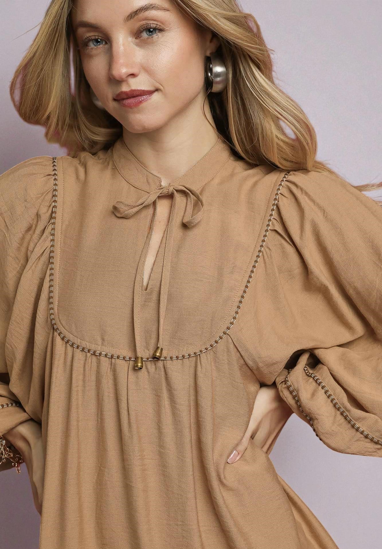 Woman wearing a brown dress with long sleeves and a tie neckline against a plain background