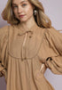 Woman wearing a brown dress with long sleeves and a tie neckline against a plain background