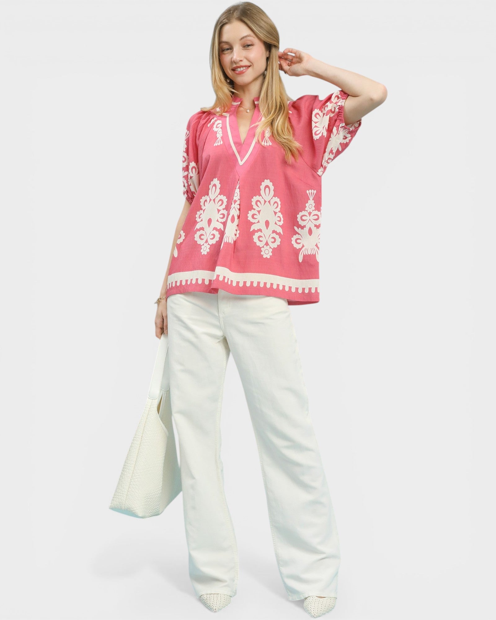 Woman wearing a pink blouse with white patterns and white pants on a white background