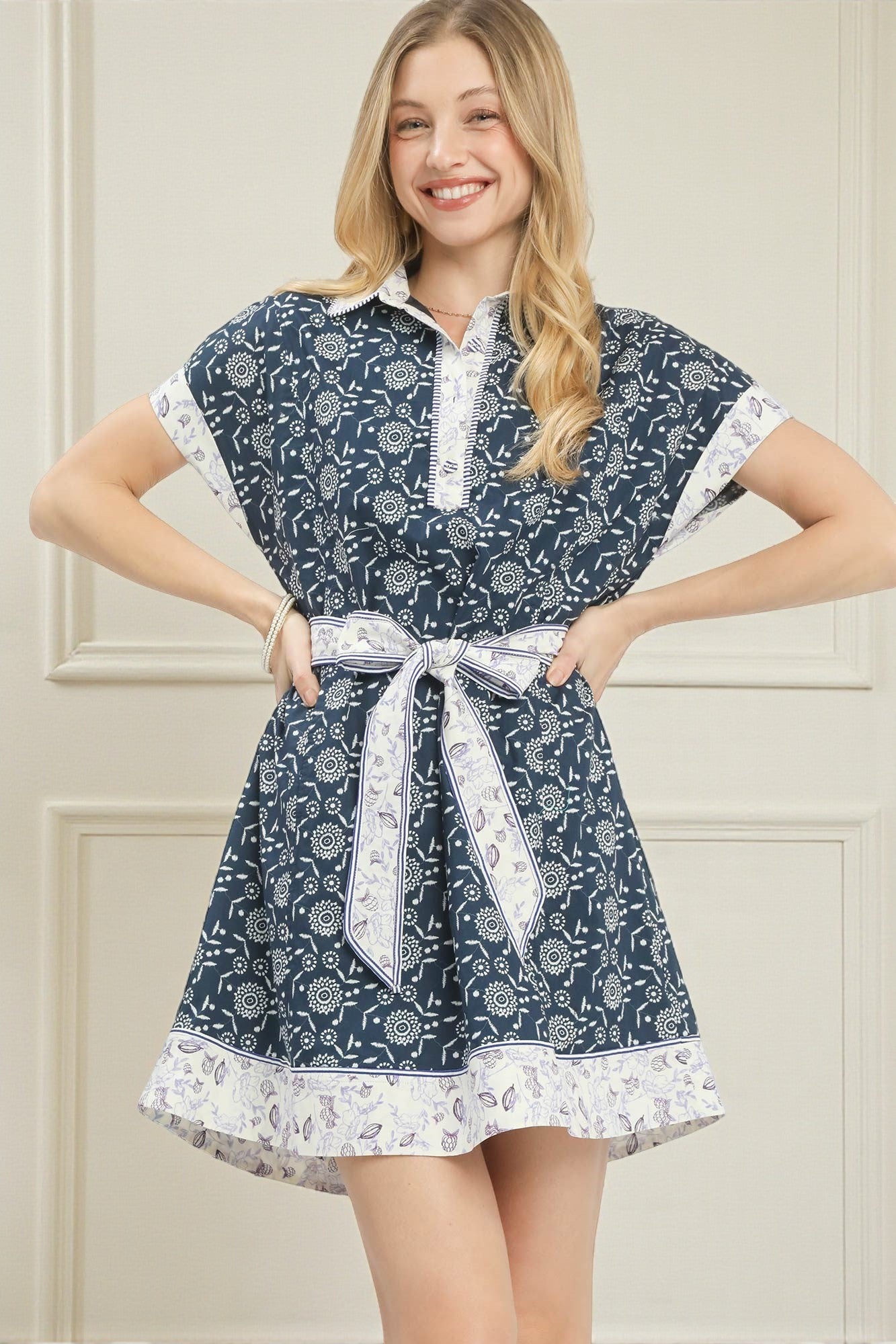 Woman wearing a blue floral dress with a white belt against a beige wall.