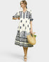 Woman wearing a white dress with black patterns, holding a woven bag and flowers, on a white background