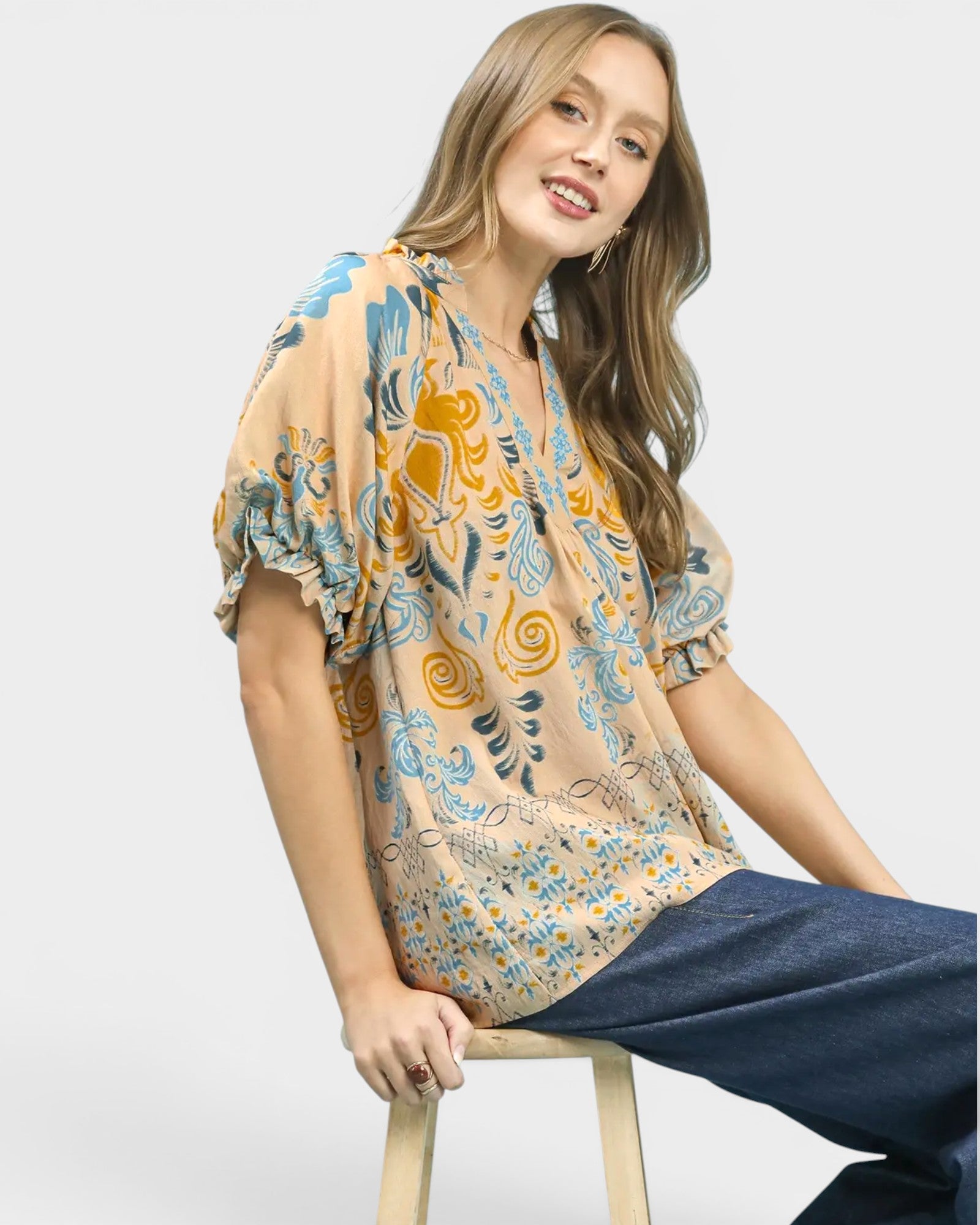 Woman wearing a patterned blouse and jeans sitting on a stool against a white background
