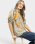 Woman wearing a patterned blouse and jeans sitting on a stool against a white background