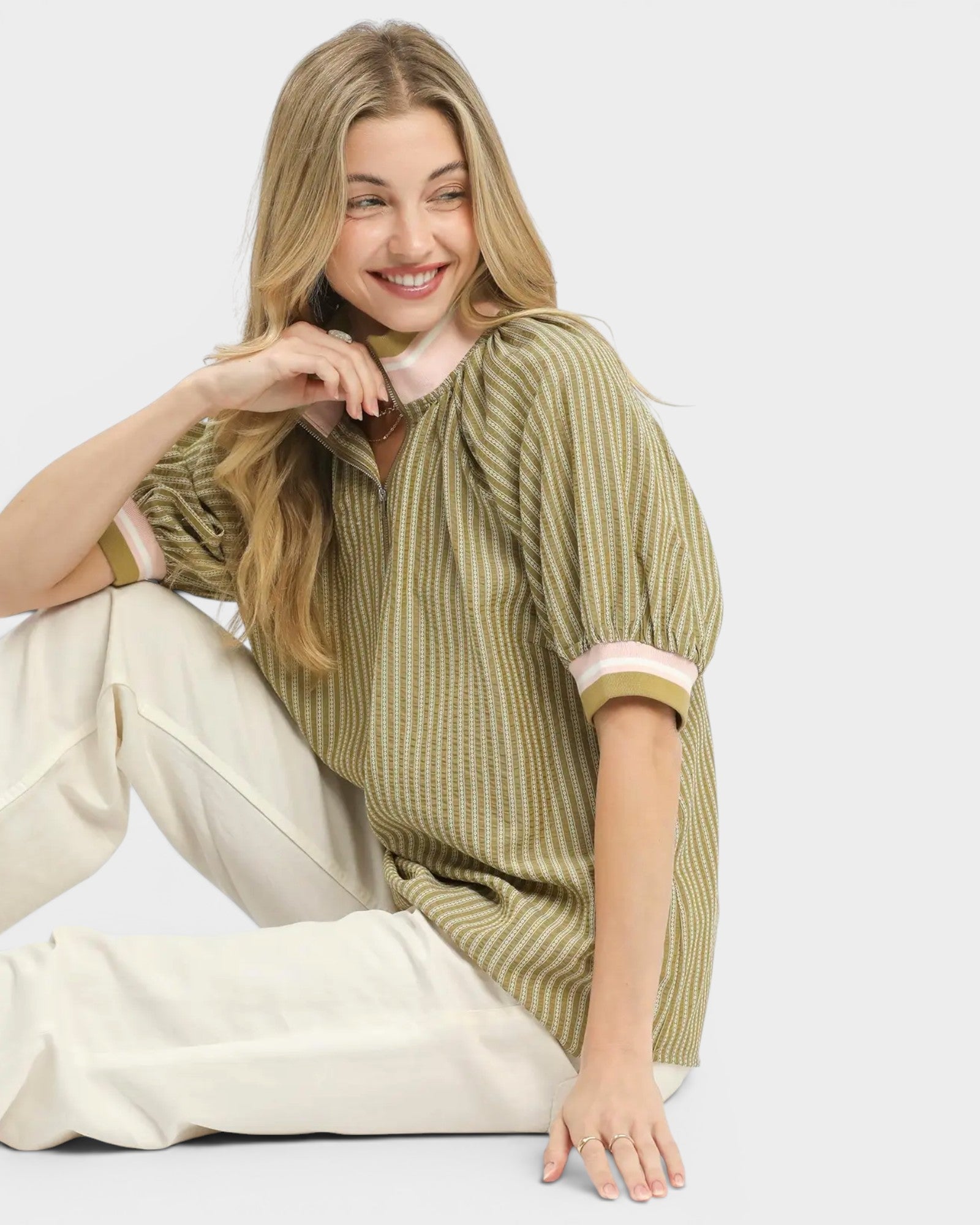 Woman wearing a green ribbed sweater and white pants sitting on a white background