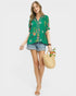 Woman wearing a green floral blouse and denim shorts, holding a straw bag with a white background