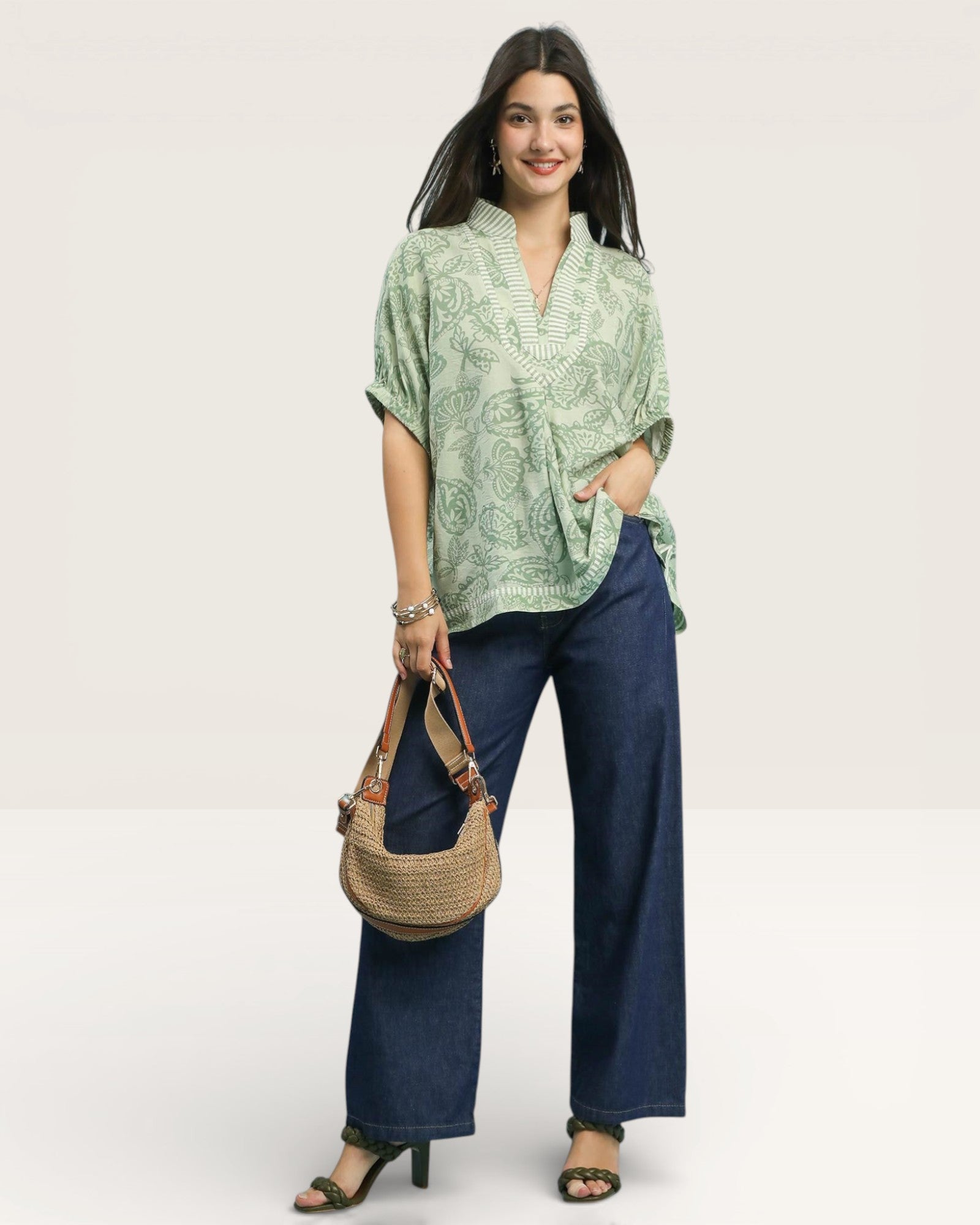 Woman wearing a green patterned top and blue pants, holding a brown handbag on a white background