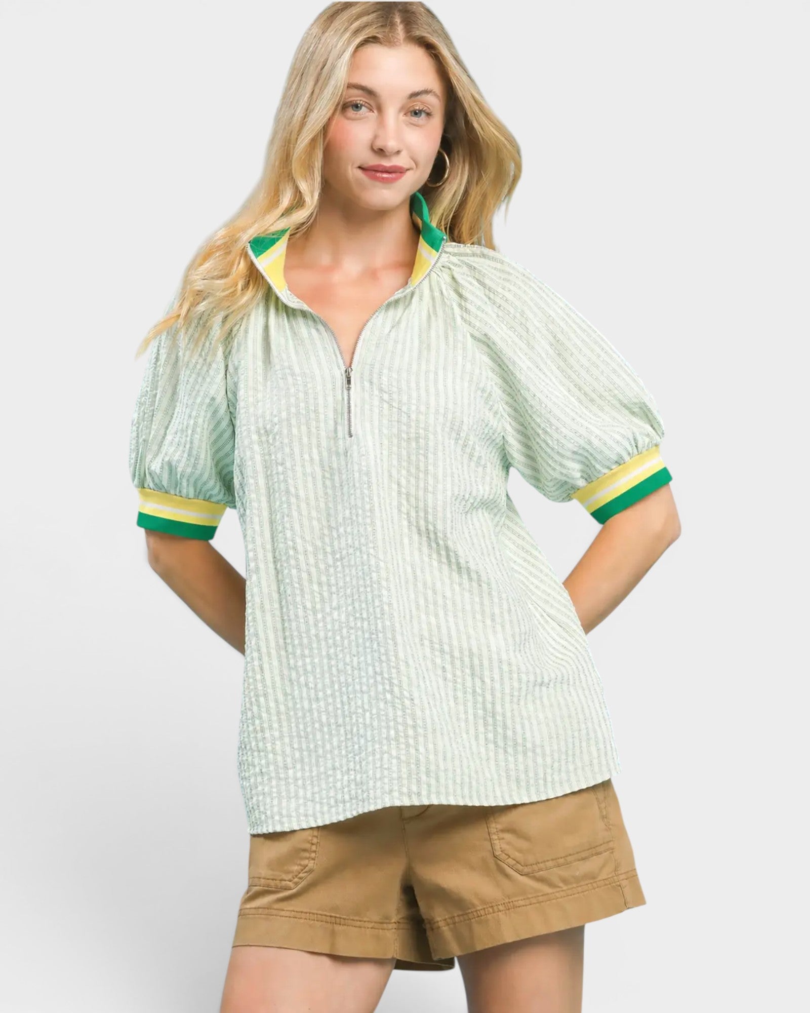 Woman wearing a light green patterned shirt with short sleeves and beige shorts on a white background