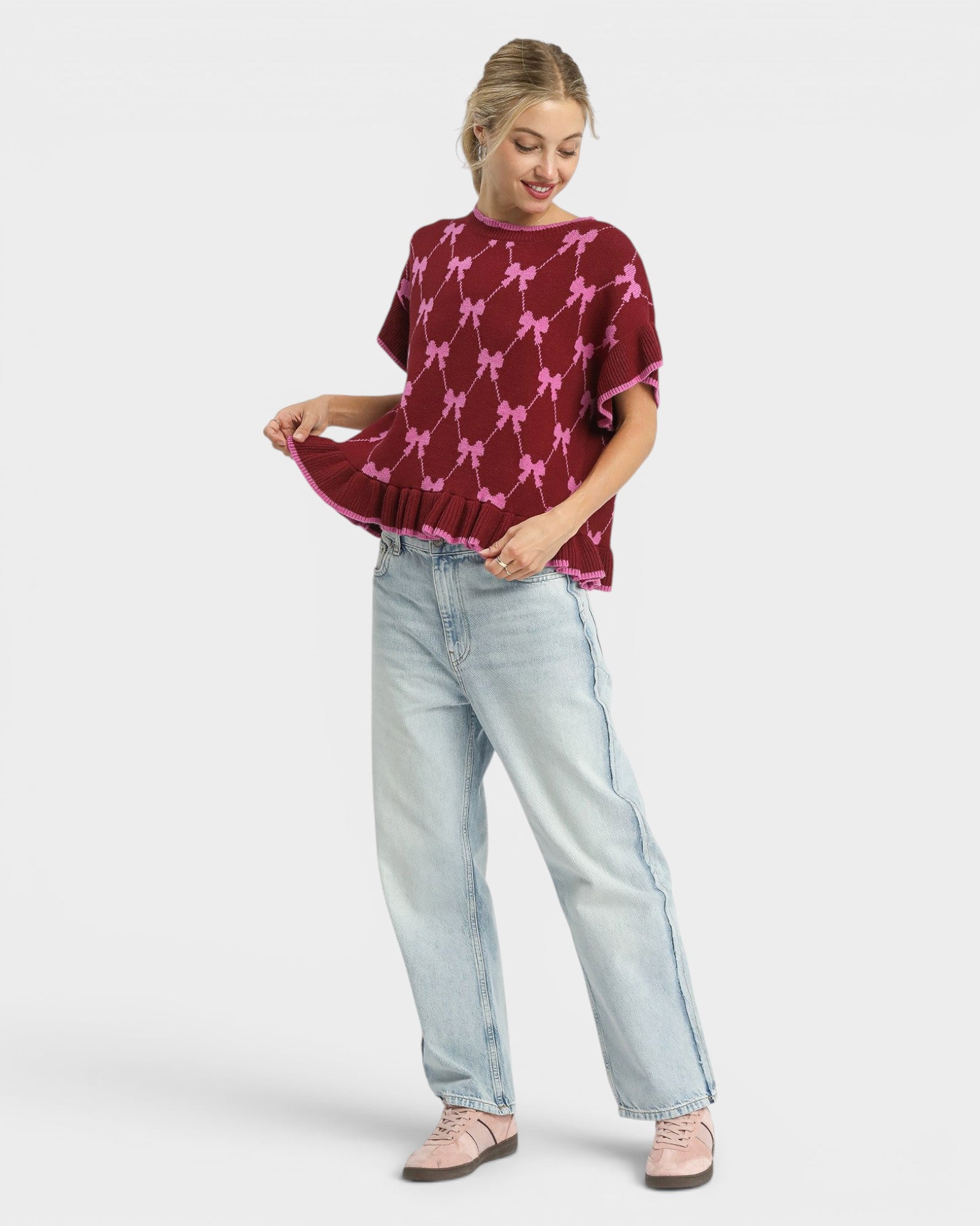 Woman wearing a burgundy sweater with pink bow pattern and light blue jeans on a white background