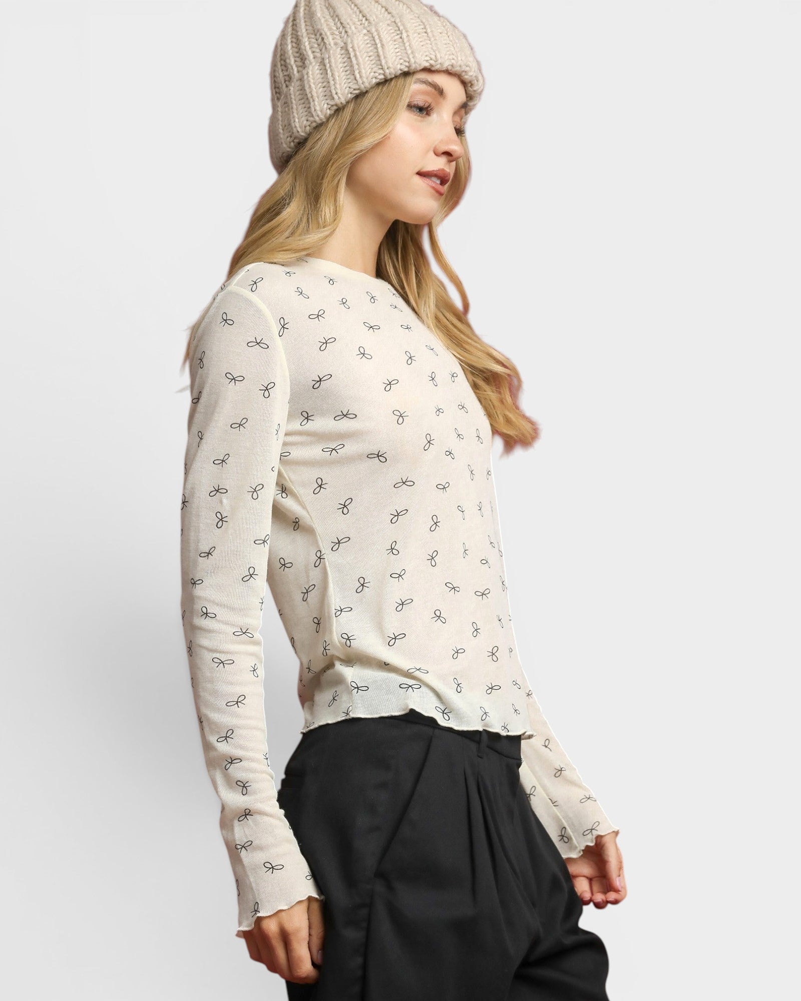 Woman wearing a white floral blouse and black pants with a beige beanie on a light gray background