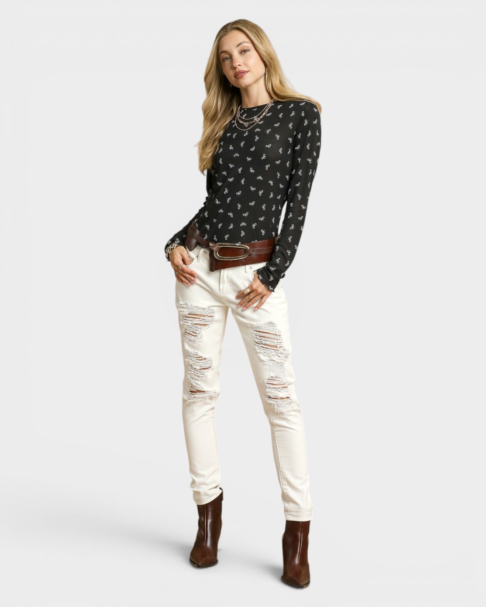 Woman wearing a black patterned top, white distressed jeans, and brown boots on a white background
