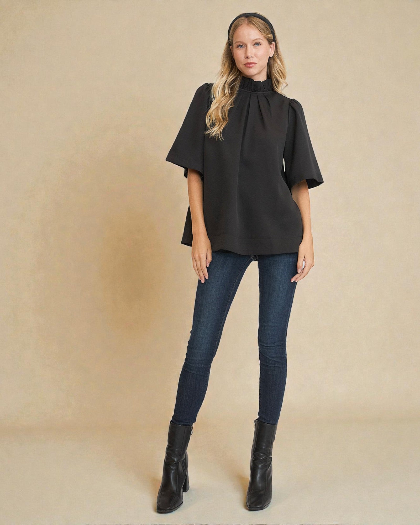 Woman wearing a black poncho, blue jeans, and black boots against a beige background